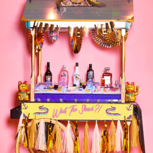 Colorful bar cart with liquor bottles and festive decor, featuring a pink background and playful accessories.